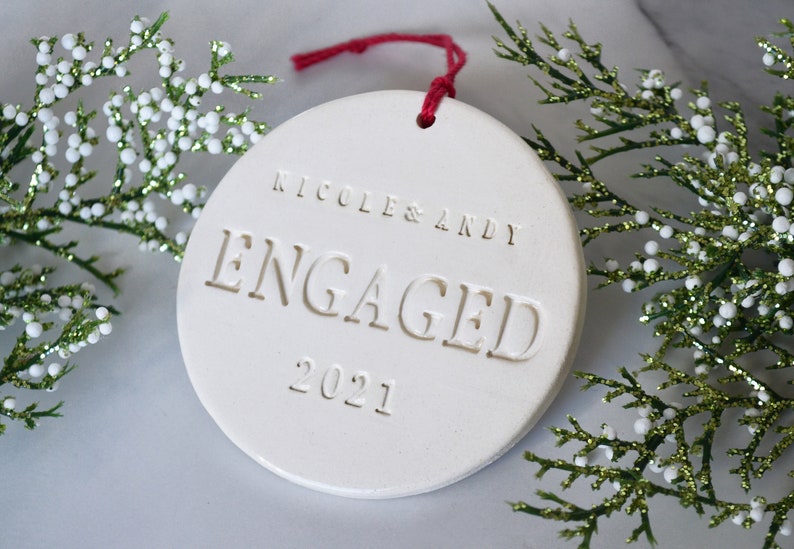 Product image of an off white gloss glazed ceramic round shaped  ornament. The front is stamped with custom text in all capital letters. The text is centered and reads Nicole & Andy, Engaged, 2021. The ornament has a red cord to hang from.