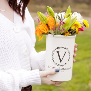 May include: White ceramic vase with a black floral wreath and the letter "V" in the center. The vase is personalized with the text "Katrina & Anthony" and the date "2022". The vase is filled with a bouquet of colorful flowers, including yellow, orange, white, and pink.
