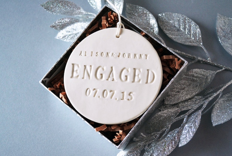 Product image of an off-white gloss-glazed ceramic round-shaped  ornament. The front is stamped with custom text in all capital letters. The text is centered and reads Alison & Johnny, Engaged, 07.07.15. The ornament has a white cord to hang from.