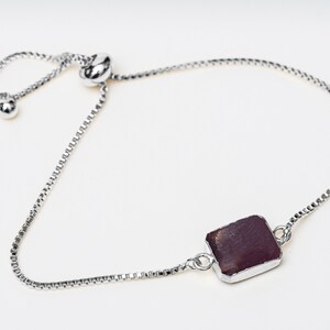 A silver chain bracelet with a dark red square gemstone pendant. The bracelet has a sliding clasp for adjustable sizing.