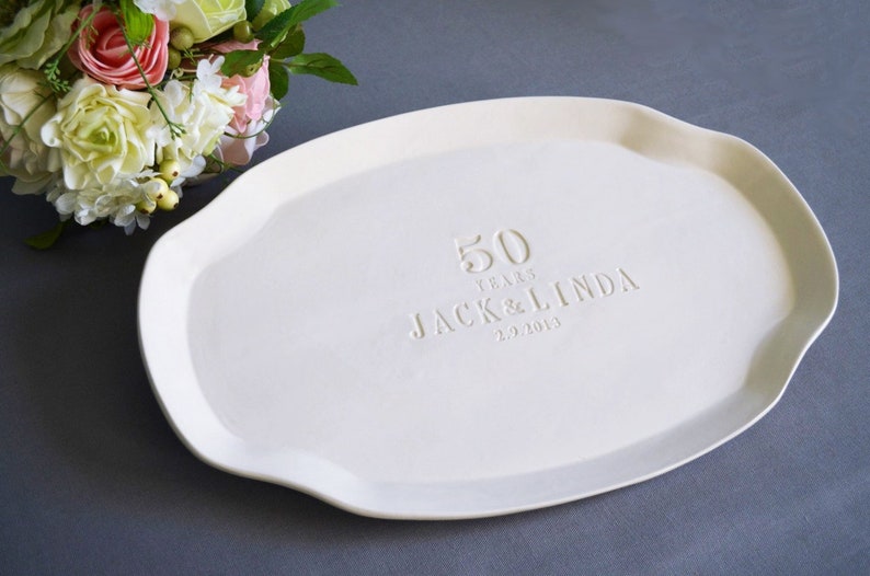 May include: An oval-shaped white ceramic serving platter featuring an embossed inscription that reads 50 YEARS JACK & LINDA 2.9.2019. The platter has a smooth, matte finish and a slightly scalloped edge, providing a clean and neutral surface.