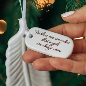 May include: White ceramic feather ornament with a white ribbon and a tag that reads "Feathers are reminders that angels are always near."
