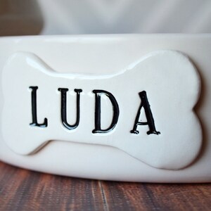 A close up image of the off-white bone shaped tile on the front of the off-white dog bowl. The pet name LUDA is stamped into the tile in all capital letters, centered, and painted with black paint. The bowl sits on a wooden table.