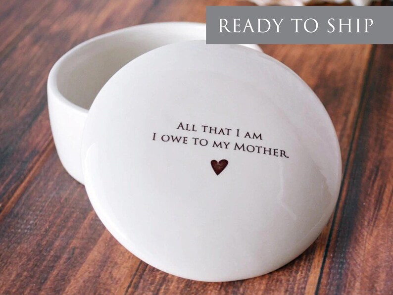 May include: White ceramic trinket box with a lid. The lid has the text "All that I am I owe to my mother." with a small heart.