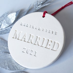 Product image of an off-white gloss-glazed ceramic round-shaped  ornament. The front is stamped with custom text in all capital letters. The text is centered and reads Marina & Steve, Married, 2021. The ornament has a red cord to hang from.