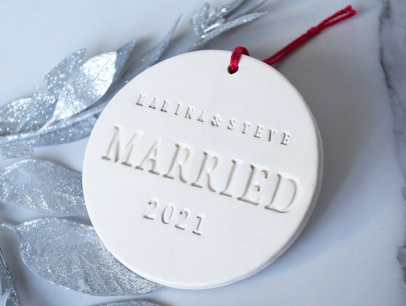 Product image of an off-white gloss-glazed ceramic round-shaped  ornament. The front is stamped with custom text in all capital letters. The text is centered and reads Marina & Steve, Married, 2021. The ornament has a red cord to hang from.