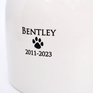 Pode incluir: Urna de cer&acirc;mica branca com letras pretas que dizem "BENTLEY 2011-2023" e uma pegada de pata preta.
