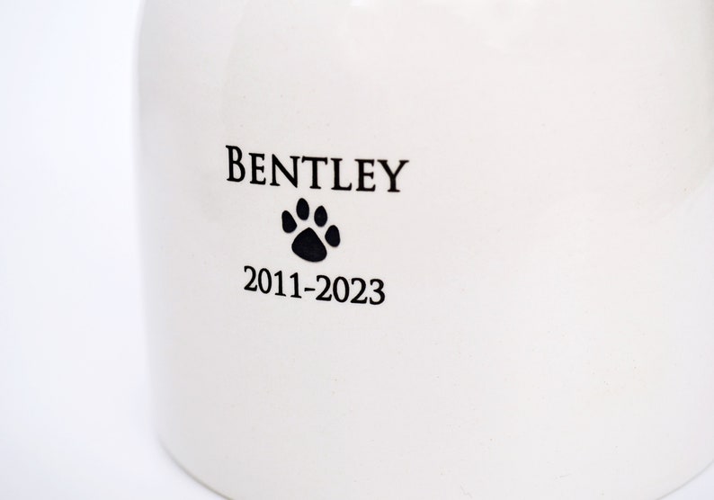 Pode incluir: Urna de cer&acirc;mica branca com letras pretas que dizem "BENTLEY 2011-2023" e uma pegada de pata preta.