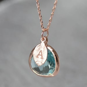 A light blue aquamarine colored transparent glass stone charm is framed in rose gold metal and hangs from a rose gold link chain. There is a small leaf shaped charm with the letter A stamped on the front, hanging in front of the aquamarine charm.