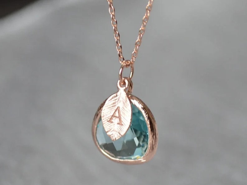 A light blue aquamarine colored transparent glass stone charm is framed in rose gold metal and hangs from a rose gold link chain. There is a small leaf shaped charm with the letter A stamped on the front, hanging in front of the aquamarine charm.
