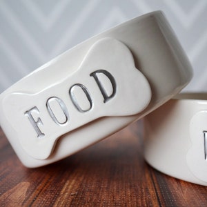 Może przedstawiać: Dwie białe ceramiczne miski dla psa z etykietami w kształcie kości. Miska po lewej stronie ma napis "FOOD", a miska po prawej stronie ma napis "WATER".