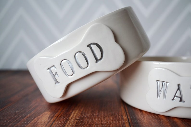 Może przedstawiać: Dwie białe ceramiczne miski dla psa z etykietami w kształcie kości. Miska po lewej stronie ma napis "FOOD", a miska po prawej stronie ma napis "WATER".