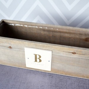 May include: A wooden planter box with a white square label that says "B" in gold lettering. The planter is made of natural wood and has a rustic look.