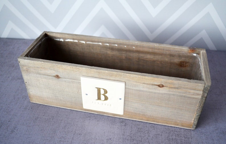May include: A wooden planter box with a white square label that says "B" in gold lettering. The planter is made of natural wood and has a rustic look.