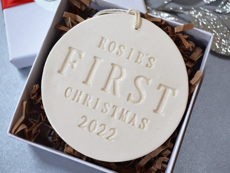 A round shaped, gloss glazed, off-white, ceramic ornament sits in a white gift box on a grey table. The ornament has a white cord to hang from and text stamped into the surface on the front. The text is in all capital letters and centered.