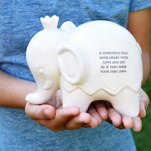 Peut inclure: Une tirelire blanche en forme d'&eacute;l&eacute;phant avec une couronne sur la t&ecirc;te. La tirelire porte une citation imprim&eacute;e sur le c&ocirc;t&eacute;. L'&eacute;l&eacute;phant est tenu dans les mains de quelqu'un sur un fond vert flou.