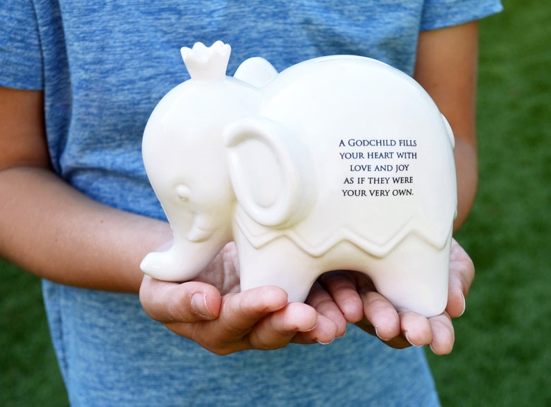 Peut inclure: Une tirelire blanche en forme d'&eacute;l&eacute;phant avec une couronne sur la t&ecirc;te. La tirelire porte une citation imprim&eacute;e sur le c&ocirc;t&eacute;. L'&eacute;l&eacute;phant est tenu dans les mains de quelqu'un sur un fond vert flou.
