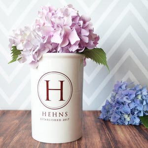 Puede incluir: Un jarr&oacute;n de cer&aacute;mica blanco con la letra "H" burdeos dentro de un c&iacute;rculo, con el nombre "HEHNS" y "ESTABLISHED 2017" debajo. El jarr&oacute;n contiene hortensias rosas. Un racimo de hortensias azules est&aacute; cerca.