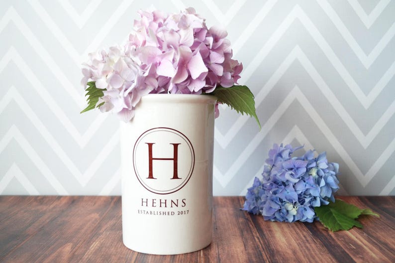 Puede incluir: Un jarr&oacute;n de cer&aacute;mica blanco con la letra "H" burdeos dentro de un c&iacute;rculo, con el nombre "HEHNS" y "ESTABLISHED 2017" debajo. El jarr&oacute;n contiene hortensias rosas. Un racimo de hortensias azules est&aacute; cerca.