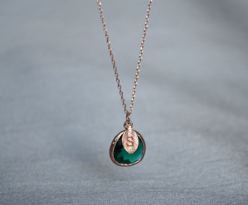 A green emerald colored transparent glass stone charm is framed in rose gold metal and hangs from a rose gold link chain. There is a small leaf shaped charm with the letter S stamped on the front, hanging in front of the emerald charm.