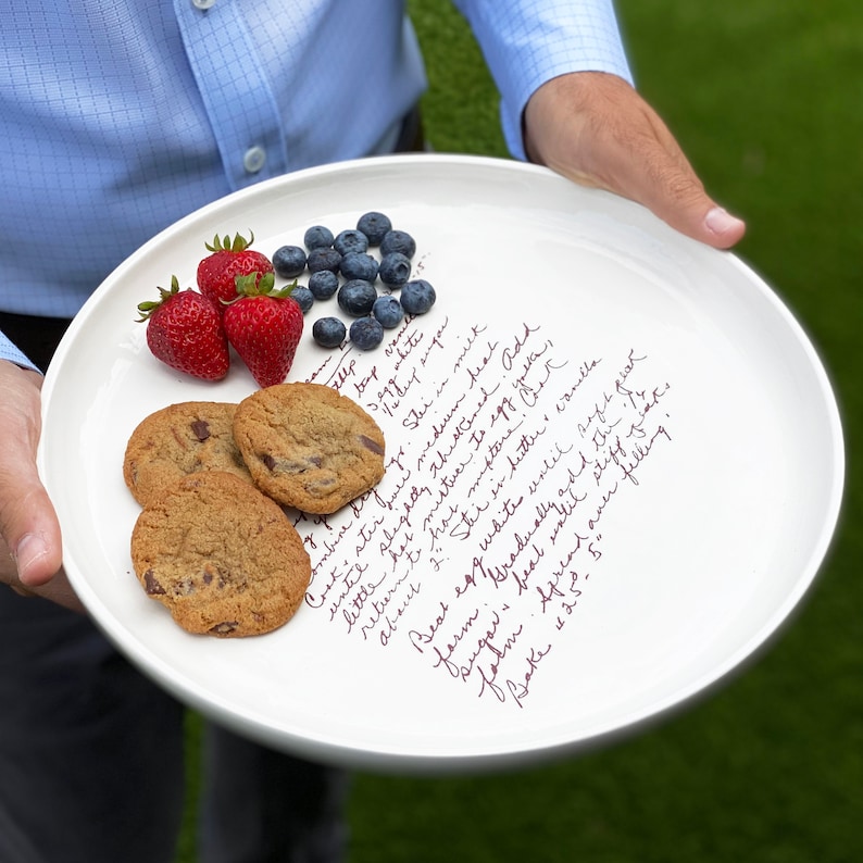 A large round serving plate is held in front of a grass background. Cookies and fruit sit on the plate for entertaining. The plate has light brown/sepia text printed on the front in a handwritten script font. The text is a custom family recipe.