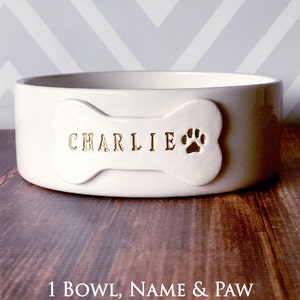 Image of the 1 bowl, name & paw option. One bowl is shown with a bone shaped tile on the front. The tile is stamped with the pet name in all capital letters with a paw print image on the right, all painted in metallic gold.