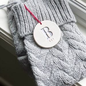 A grey knitted holiday stocking with off-white, round, ceramic ornament hanging from red cord. A large letter B is stamped into the ornament and painted in metallic silver. The stocking is folded on a windowsill with the ornament resting on top.