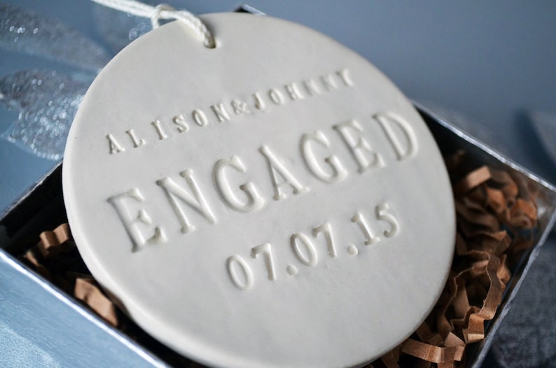 Product image of an off white gloss glazed ceramic round shaped ornament. The front is stamped with custom text in all capital letters. The text is centered and reads Alison & Johnny, Engaged, 07.07.15. The ornament has a white cord to hang from.