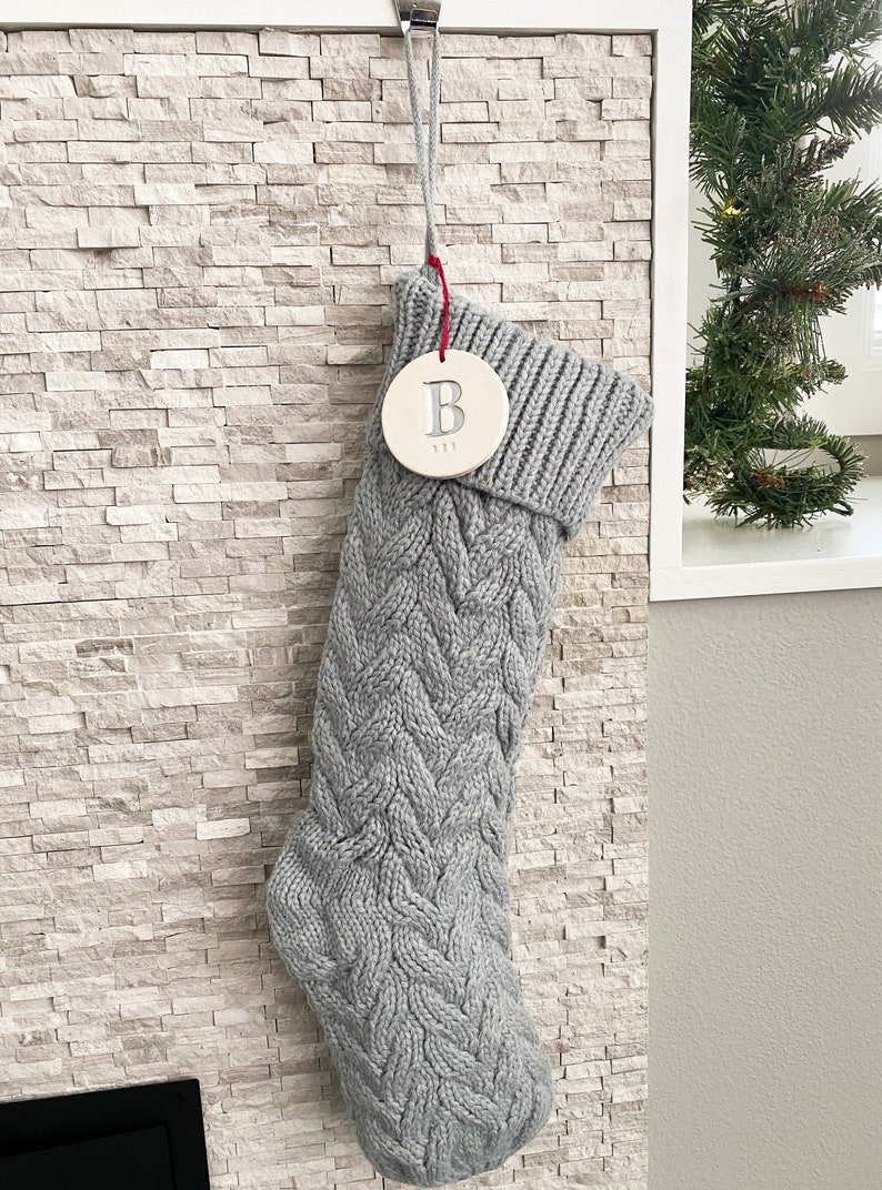 A grey knitted holiday stocking with off-white, round, ceramic ornament hanging from red cord. A large letter B is stamped into the ornament and painted in metallic silver. The name "BEN" is stamped below the initial in smaller text.