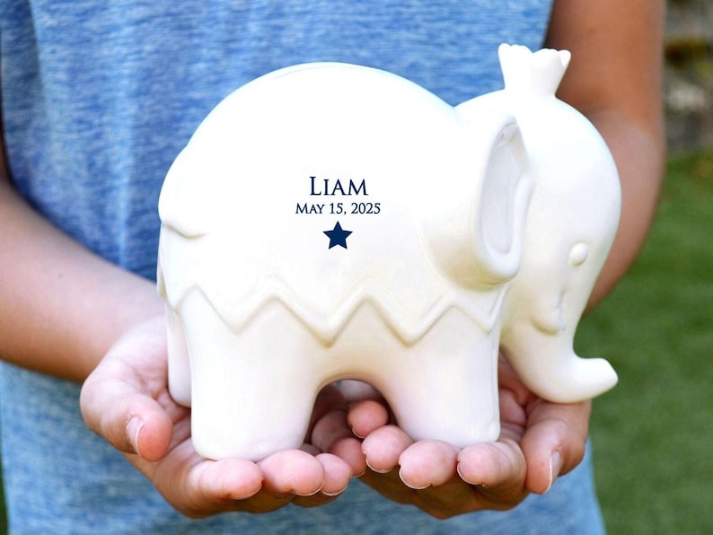 Peut inclure: Figurine d'&eacute;l&eacute;phant en c&eacute;ramique blanche avec un motif en zigzag sur le corps. Le texte "LIAM MAY 15, 2025" est imprim&eacute; en bleu marine. L'&eacute;l&eacute;phant est tenu dans les mains.