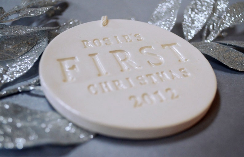 A flat round shaped, gloss glazed, off-white, ceramic ornament viewed from the side, sits on a grey table. The ornament is thin in thickness with glaze and stamped text on the surface.