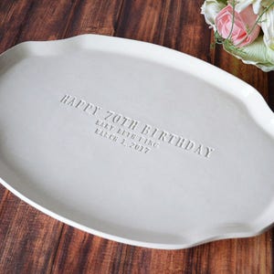 A white ceramic serving platter with a gently scalloped edge. The platter is inscribed with "HAPPY 70TH BIRTHDAY" and the name "Mary Beth Ying" and the date "March 1, 2017". A bouquet of flowers is in the background.