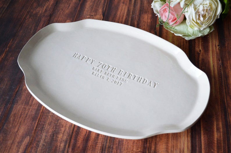 A white ceramic serving platter with a gently scalloped edge. The platter is inscribed with "HAPPY 70TH BIRTHDAY" and the name "Mary Beth Ying" and the date "March 1, 2017". A bouquet of flowers is in the background.