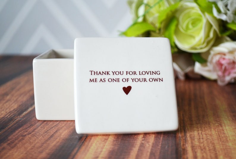 May include: A white square ceramic box with a lid. The lid has the text "THANK YOU FOR LOVING ME AS ONE OF YOUR OWN" in burgundy, with a burgundy heart below. The box is on a wooden surface.