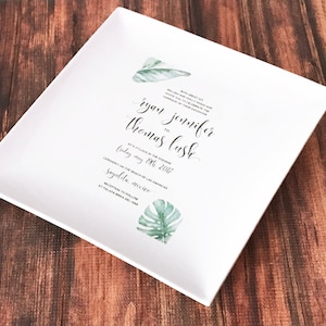 May include: White square ceramic plate with a wedding invitation printed on it. The invitation features a tropical leaf design and the text "With great joy we card you to celebrate the marriage of Ryan Thomas to Jennifer Lush at 4 o'clock in the evening Friday, May 19th, 2017 Ceremony on the beach of Las Palmas Sayulita, Mexico Reception to follow at Palapa Bonita del Mar"