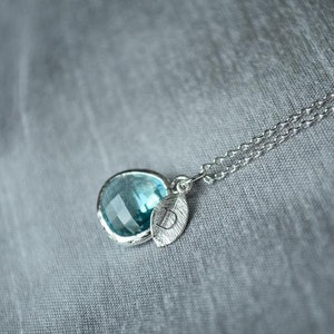 A light blue aquamarine colored transparent glass stone charm is framed in silver metal and hangs from a silver link chain. There is a small leaf shaped charm with the letter D stamped on the front, hanging in front of the aquamarine charm.