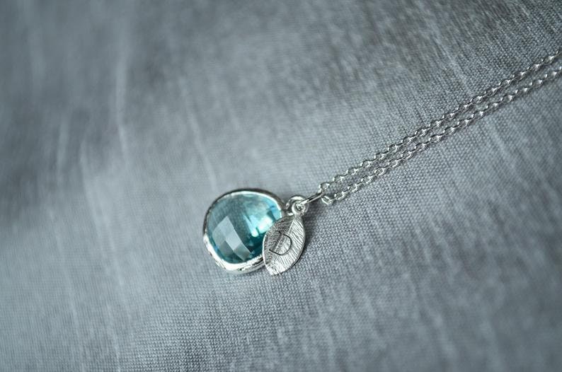 A light blue aquamarine colored transparent glass stone charm is framed in silver metal and hangs from a silver link chain. There is a small leaf shaped charm with the letter D stamped on the front, hanging in front of the aquamarine charm.
