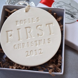 A round shaped, gloss glazed, off-white, ceramic ornament sits in a white gift box on a grey table. The ornament has a white cord to hang from and text stamped into the surface on the front. The text is in all capital letters and centered.