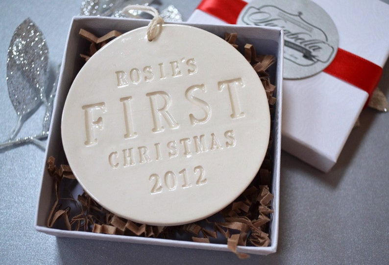 A round shaped, gloss glazed, off-white, ceramic ornament sits in a white gift box on a grey table. The ornament has a white cord to hang from and text stamped into the surface on the front. The text is in all capital letters and centered.