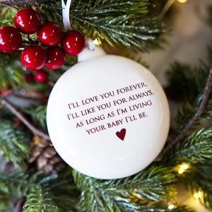 May include: A round white ceramic Christmas ornament featuring the printed text: I'LL LOVE YOU FOREVER, I'LL LIKE YOU FOR ALWAYS, AS LONG AS I'M LIVING YOUR BABY I'LL BE, followed by a small dark red heart. The ornament hangs from a white ribbon.