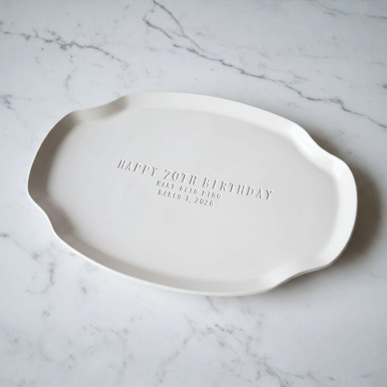 A white ceramic serving platter with a gently scalloped edge. The platter is inscribed with "HAPPY 70TH BIRTHDAY" and the name "Mary Beth Ping" and the date "March 1, 2026".