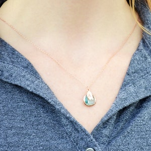 The necklace is shown on a model wearing a grey button down shirt. A rose gold metal chain with aquamarine charm and leaf charm hangs around the model's neck. The aquamarine is framed in rose gold and the leaf on top is stamped with an initial.