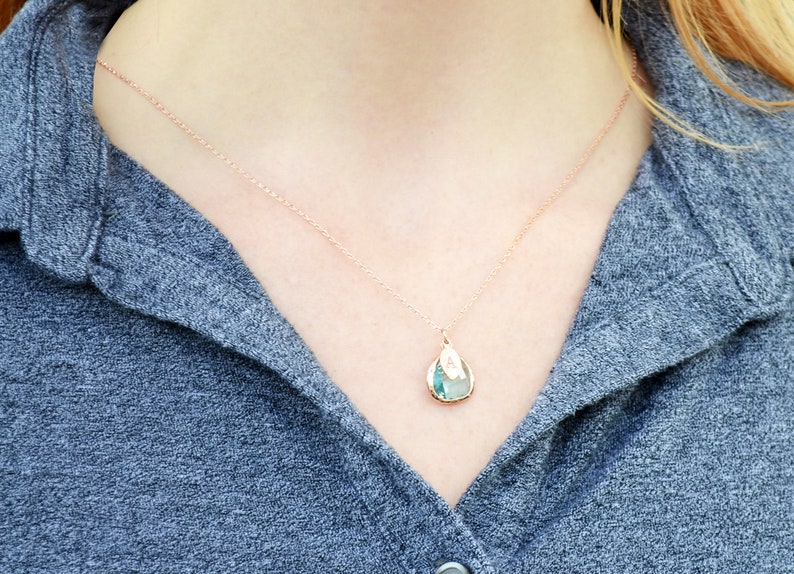 May include: A delicate rose gold necklace with a teardrop pendant. The pendant features a light blue and green stone with a gold-colored border. The necklace is worn against a gray shirt.