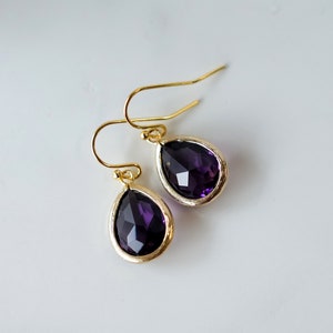 K&ouml;nnte beinhalten: Ein Paar goldene Tropfenohrringe mit violetten facettierten Steinen.