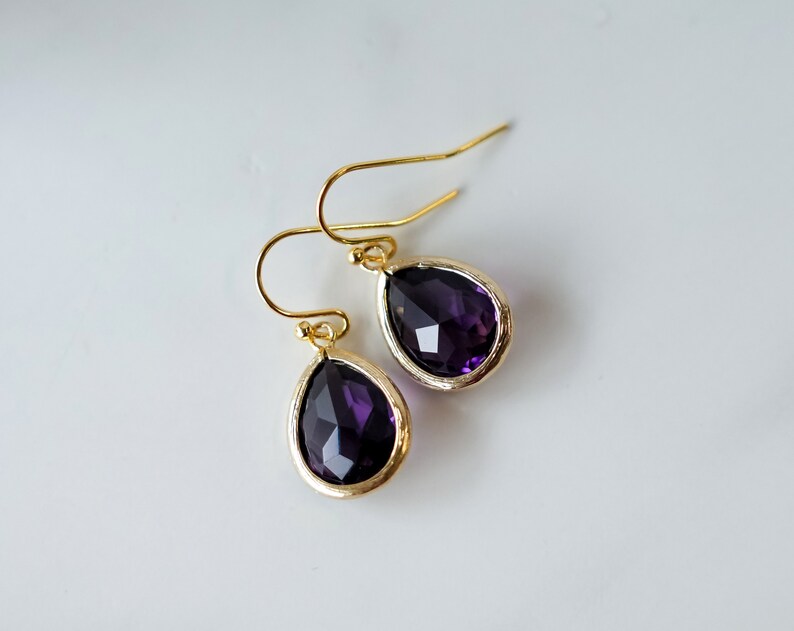K&ouml;nnte beinhalten: Ein Paar goldene Tropfenohrringe mit violetten facettierten Steinen.