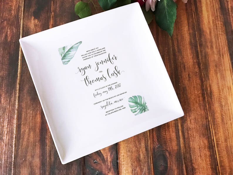 May include: White ceramic square plate with a wedding invitation printed on it. The invitation features a tropical theme with green leaves and the text "With great joy, William and Holly Fraser invite you to celebrate the marriage of their daughter, ryan jennifer to thomas lush, at 5 o'clock in the evening, Friday, May 19th, 2017, Ceremony on the beach of Las Hadas, Sayulita, Mexico, Reception to follow at Palapa Bar, Sayulita, Mex"