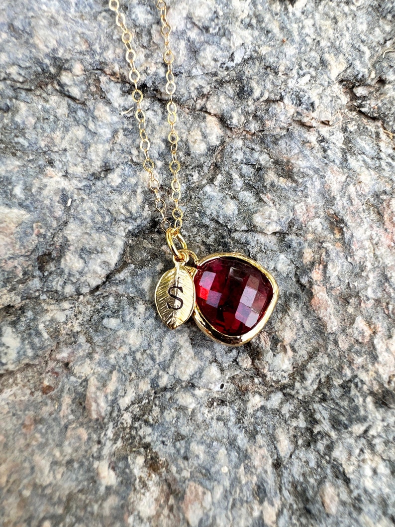 May include: A gold chain necklace with a red heart-shaped pendant and a small gold leaf charm with the letter 'S' engraved on it.