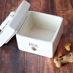 K&ouml;nnte beinhalten: Wei&szlig;e Keramikdose mit Deckel, personalisiert mit dem Namen "Rosie" und einem Pfotenabdruck. Die Dose ist leer und steht auf einer Holzoberfl&auml;che.