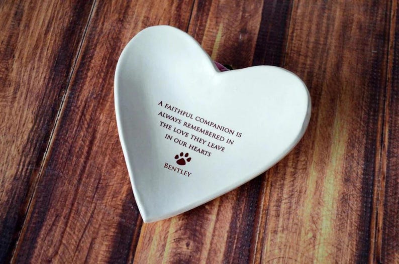 May include: White ceramic heart-shaped dish with a quote about a faithful companion and the love they leave in our hearts. The dish is personalized with the name "Bentley" and a paw print.