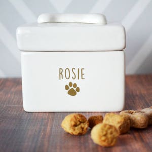 White ceramic dog treat jar with a paw print and the name "Rosie" on the front. The jar has a lid with a bone-shaped handle. There are dog treats on a wooden surface in front of the jar.